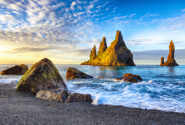Rock formations rise majestically from the ocean, with waves gently lapping at the rocky shoreline under a vibrant blue sky and scattered clouds, illuminated by the warm glow of sunset.